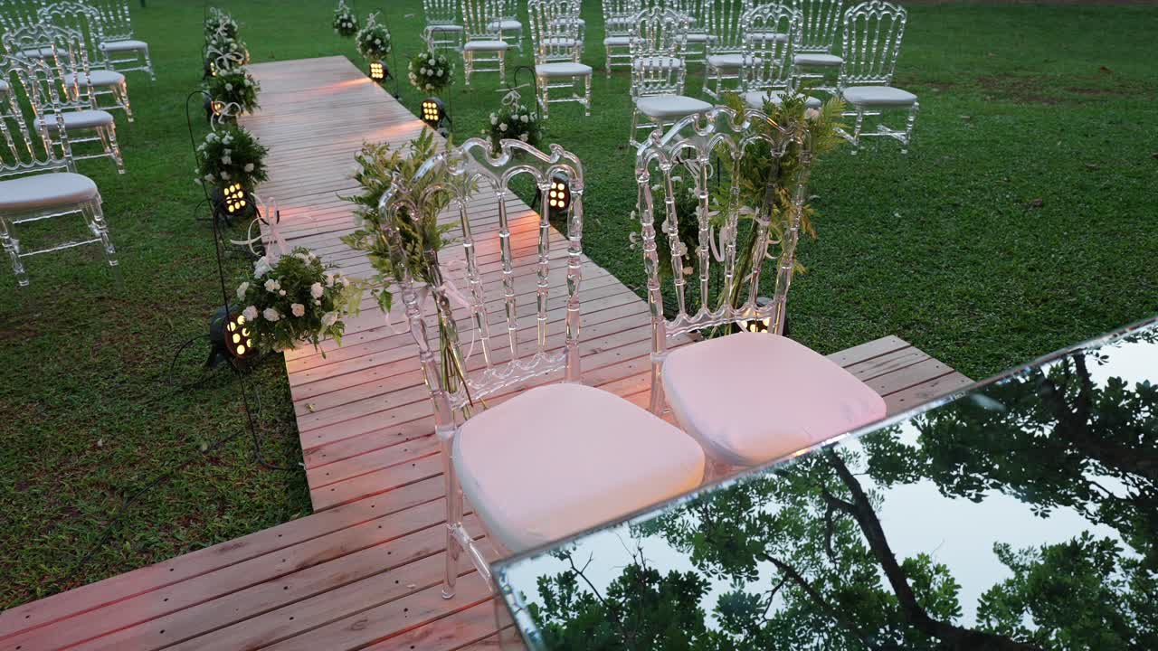 Navata nuziale simmetrica con sedie trasparenti, decorazioni floreali e illuminazione a terra soffusa sull'erba, creando un'elegante ambientazione per cerimonia all'aperto, con la telecamera che ruota e svela