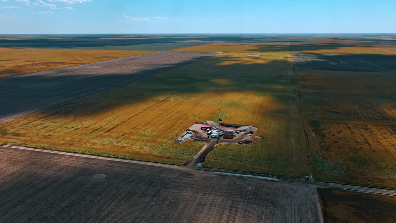 Immense field with a little site where gas is produced. High derrick towering above the cars standing at the location. Aerial perspective.
