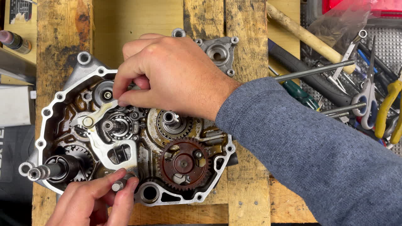 Nærbillede af mekaniker, der monterer gearstangens aksel med skiftearmen i højre side af en åben scooter-motor, og demonstrerer præcis gør-det-selv-samling på et hjemmeværksted