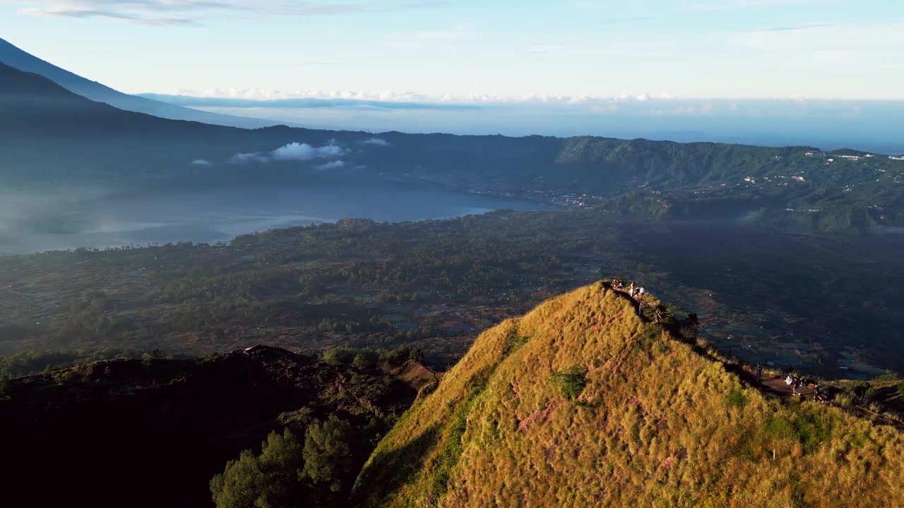Caminhantes alcançando o pico da montanha acima de um lago de cratera e um vasto vale vulcânico durante a suave luz do início da manhã em Bali, Indonésia, em imagens aéreas de drone.