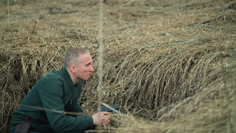 a close view of a man crouching in dry grass, holding a handgun, wearing a green jacket, looking around cautiously