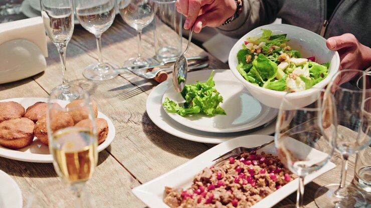Nærbillede af hænder, der serverer en skål frisk grøn salat på et solbeskinnet træbord dækket med georgiske forretter og mousserende vin