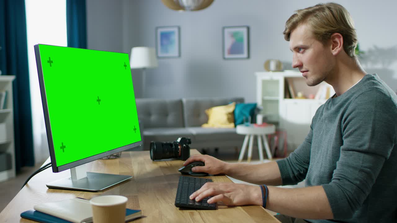 잘생긴 젊은 사진작가는 친근한 아파트에서 책상에 앉아있는 동안 녹색 모형 화면 개인 컴퓨터에서 일합니다.