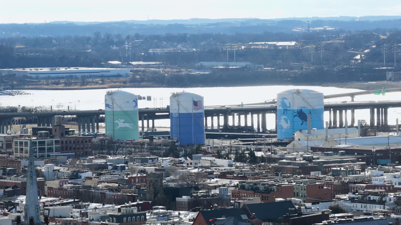 Veduta aerea del porto di Baltimora con silos industriali dipinti, ponte autostradale sull'acqua e fitti tetti urbani lungo il quartiere portuale sotto la luce chiara del giorno. Inquadratura ampia.