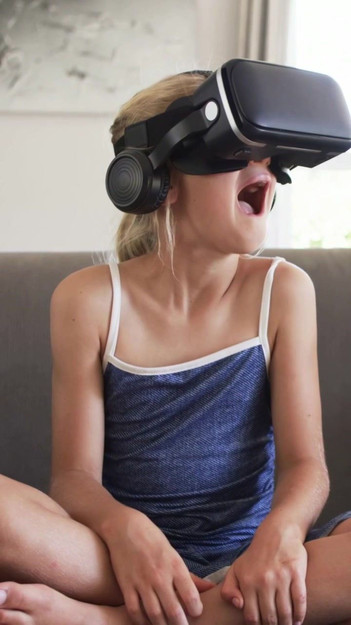 Video vertical: Chica con gafas de realidad virtual reacciona, girando, jadeando, sonriendo y extendiendo la mano en un sofá, vestida con una blusa azul.