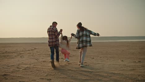 Rear-view-of-a-happy-brunette-girl-in-a-green-checkered-shirt,-together-with-her-husband,-a-middle-aged-man-with-gray-hair-in-a-checkered-shirt,-running-along-the-deserted-seashore-and-lifting-up-their-little-daughter,-a-blonde-girl-in-a-white-jacket-and-pink-pants,-during-their-vacation-outside-the