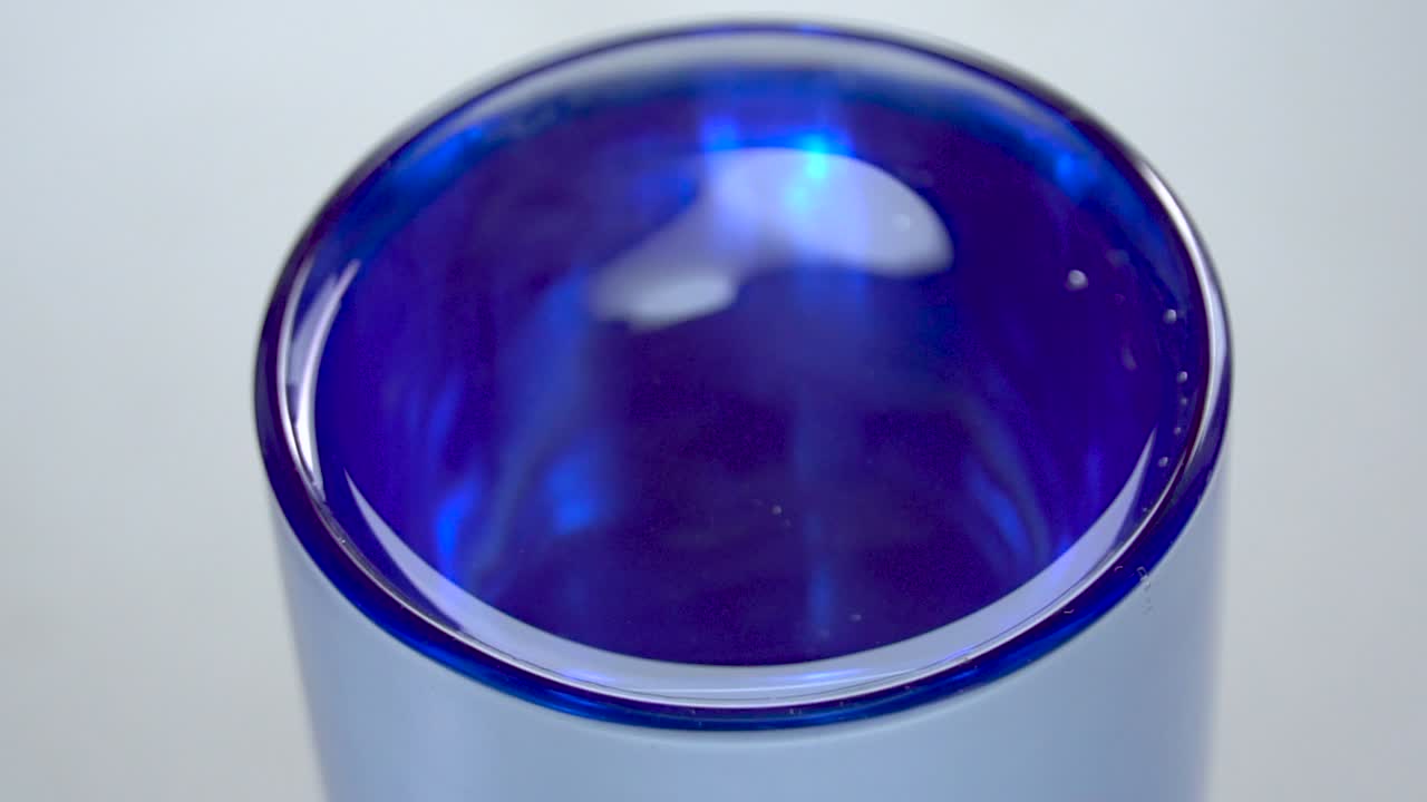 Selective focus close up view of water droplet falling into transparent glass producing splash and ripples in slow motion.