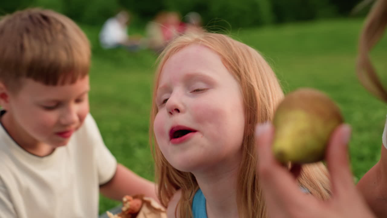 young girl chewing snack with guardian hand on shoulder, opening teeth playfully, park grass background, warm light, casual family moment showing trust and fun during outdoor picnic