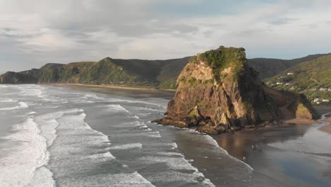 SLOWMO---Breathtaking-aerial-drone-shot-of-Lion-Rock-on-Piha-Beach-with-surfing-waves-during-sunset