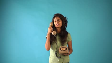 A-young-Indian-girl-in-orange-frock-standing-with-old-vintage-telephone-talking-smiling-in-an-isolated-blue-background