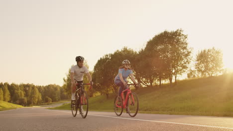 Un-Hombre-Y-Una-Mujer-En-Bicicleta-Recorren-Juntos-La-Carretera-Al-Atardecer-En-Cámara-Lenta.-La-Pareja-Viaja-En-Bicicleta.-Cascos-De-Ciclismo-Deportivos.