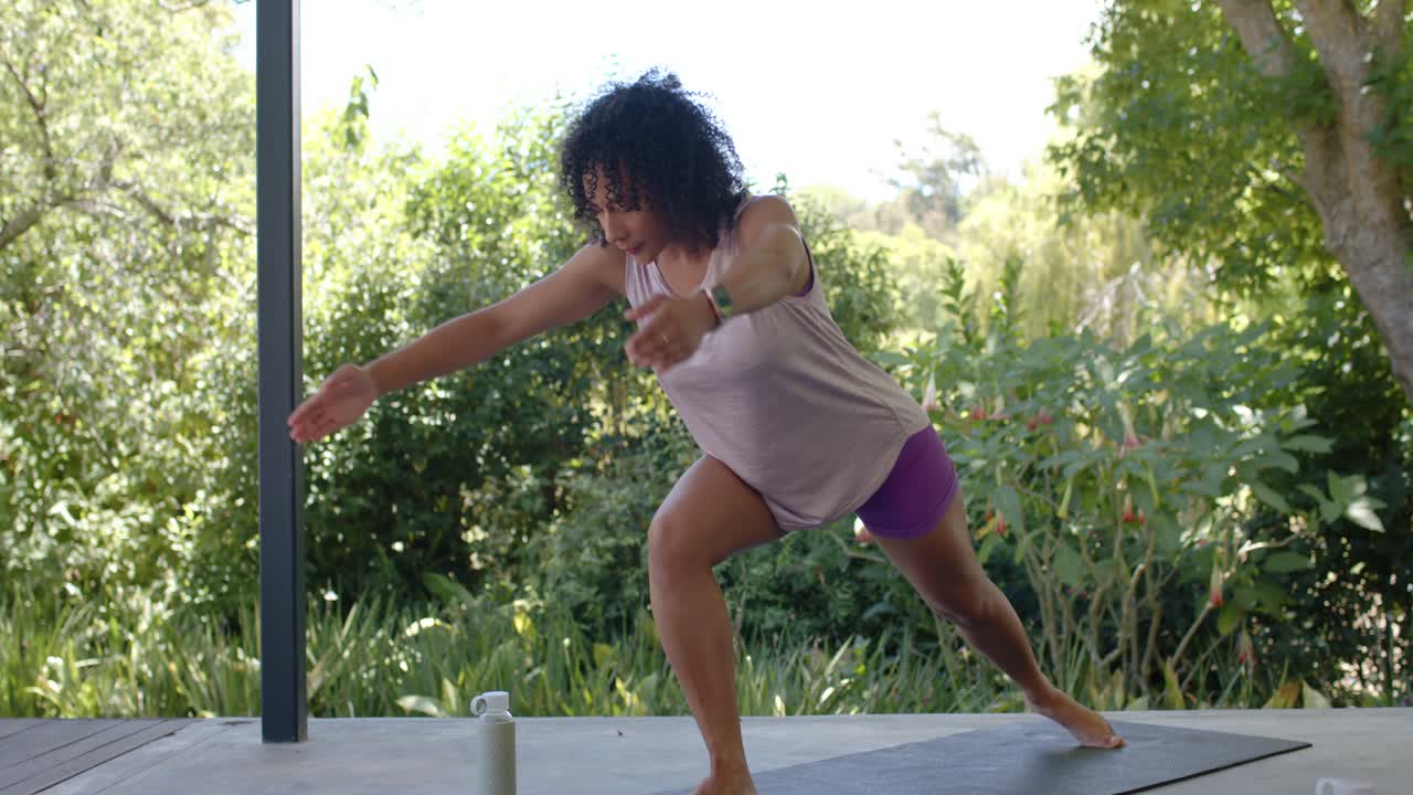 Adult African American woman following cue doing yoga on mat at patio, reaching bottle for balance