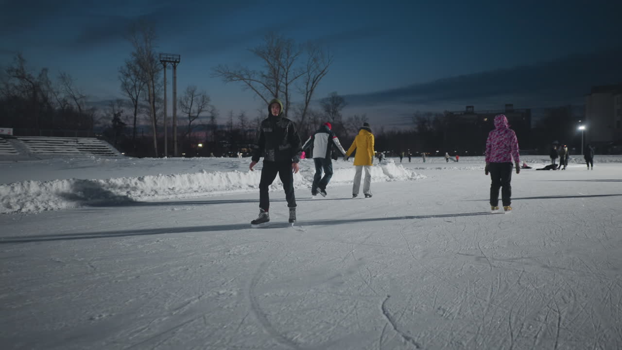 skater gliding skillfully on wide outdoor ice rink at night surrounded by other skaters illuminated by intense stadium lights with visible building structures and snowy banks