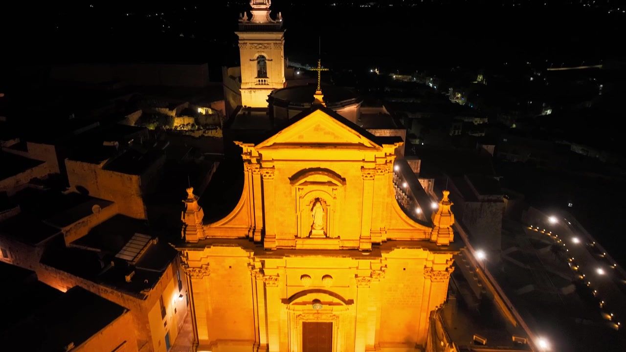 Ripresa cinematografica con drone di una facciata storica di una chiesa maltese che risplende di calde luci arancioni di notte, mostrando l'architettura mediterranea.