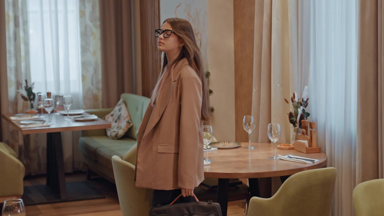 Joven mujer en una cafetería boutique se sienta con una maleta en una mesa puesta, escanea la habitación, luego se levanta y se va, iluminación cálida, sillas verdes acolchadas, mesa de madera con platos y vasos, cortinas suaves y planta en maceta