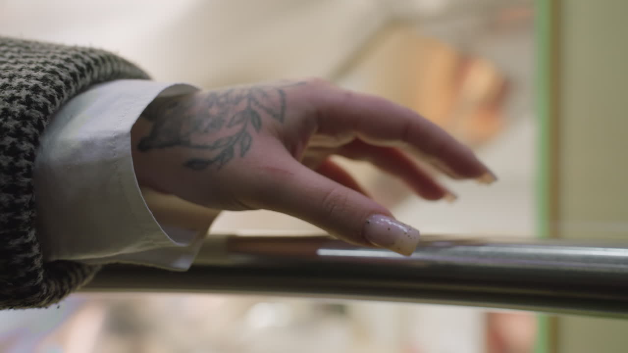 close up of tattooed hand with manicured nails gently tapping polished iron elevator rail with blurred background visible through glass panel inside descending elevator