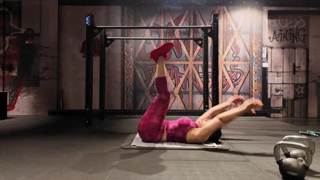 wide shot of an athletic woman in pink activewear strengthening her core by doing toe touch crunches on an exercise mat in an industrial-style gym