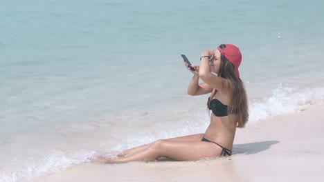 long dark haired woman sits on beach water edge with phone