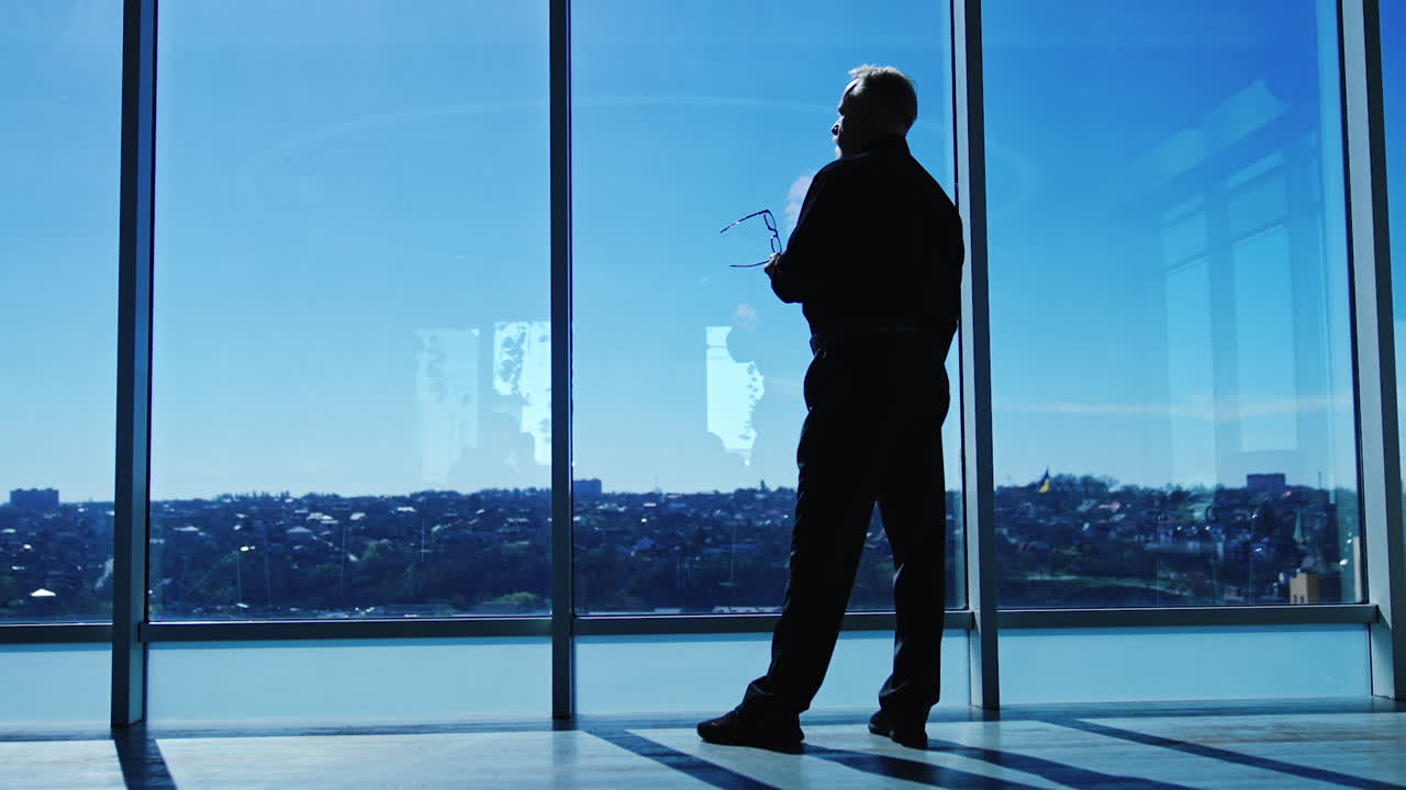 Dark silhouette of a man standing at the panoramic window in office on sunny day. Businessman thinking over issues taking off his glasses looks at cityscape. Low angle view.