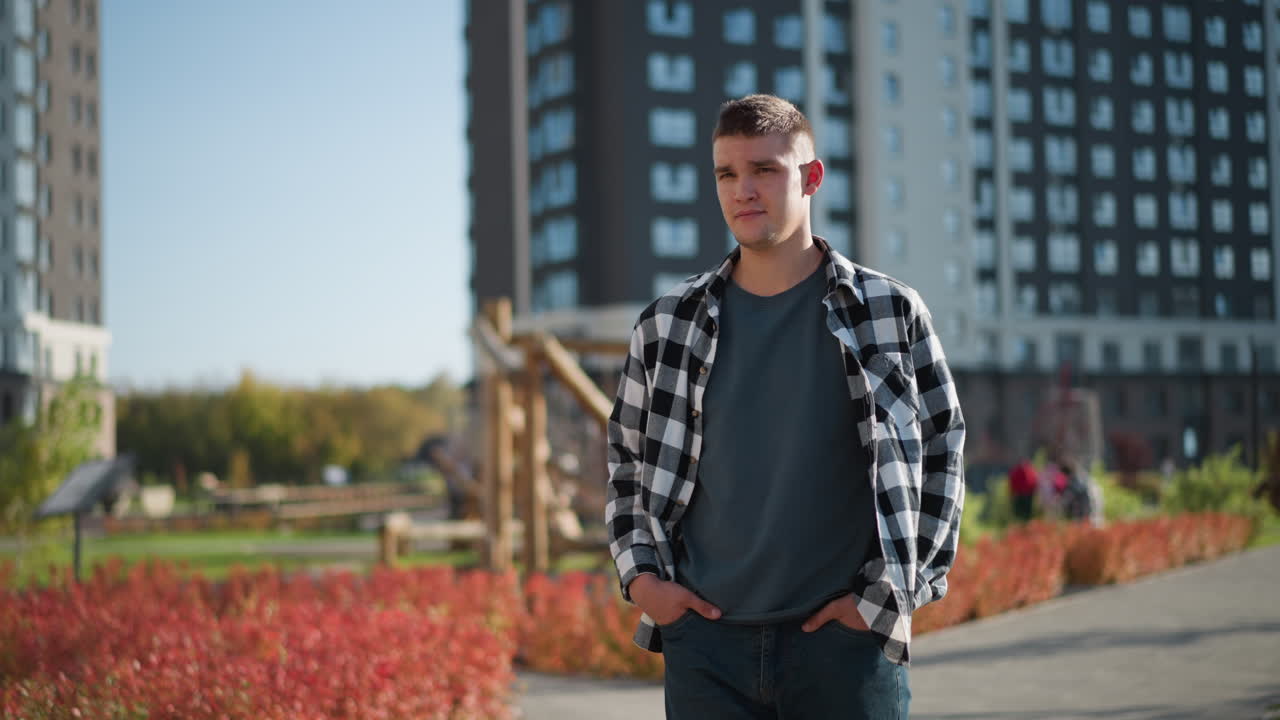 Young man wearing checkered shirt walks with hands in pockets on paved path during sunny day in modern residential park with blurred children playing in background near wooden playground structure