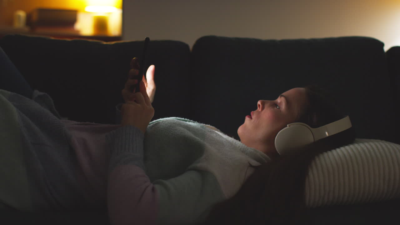 Woman Wearing Wireless Headphones Lying On Sofa At Home At Night Streaming Or Looking At Online Content On Mobile Phone 3