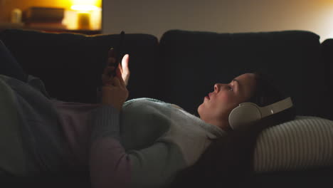 woman wearing wireless headphones lying on sofa at home at night streaming or looking at online content on mobile phone 3