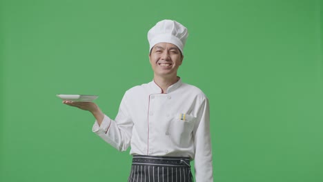 chef asiático sosteniendo un plato y sonriendo a la cámara mientras está de pie en el estudio de fondo de la pantalla verde