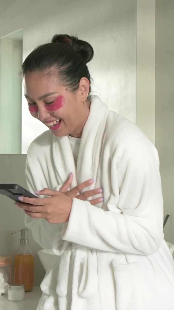 Video verticale: Donna asiatica in accappatoio che prende prodotti per la cura della pelle, applica patch occhi rosa al lavandino e usa il telefono.