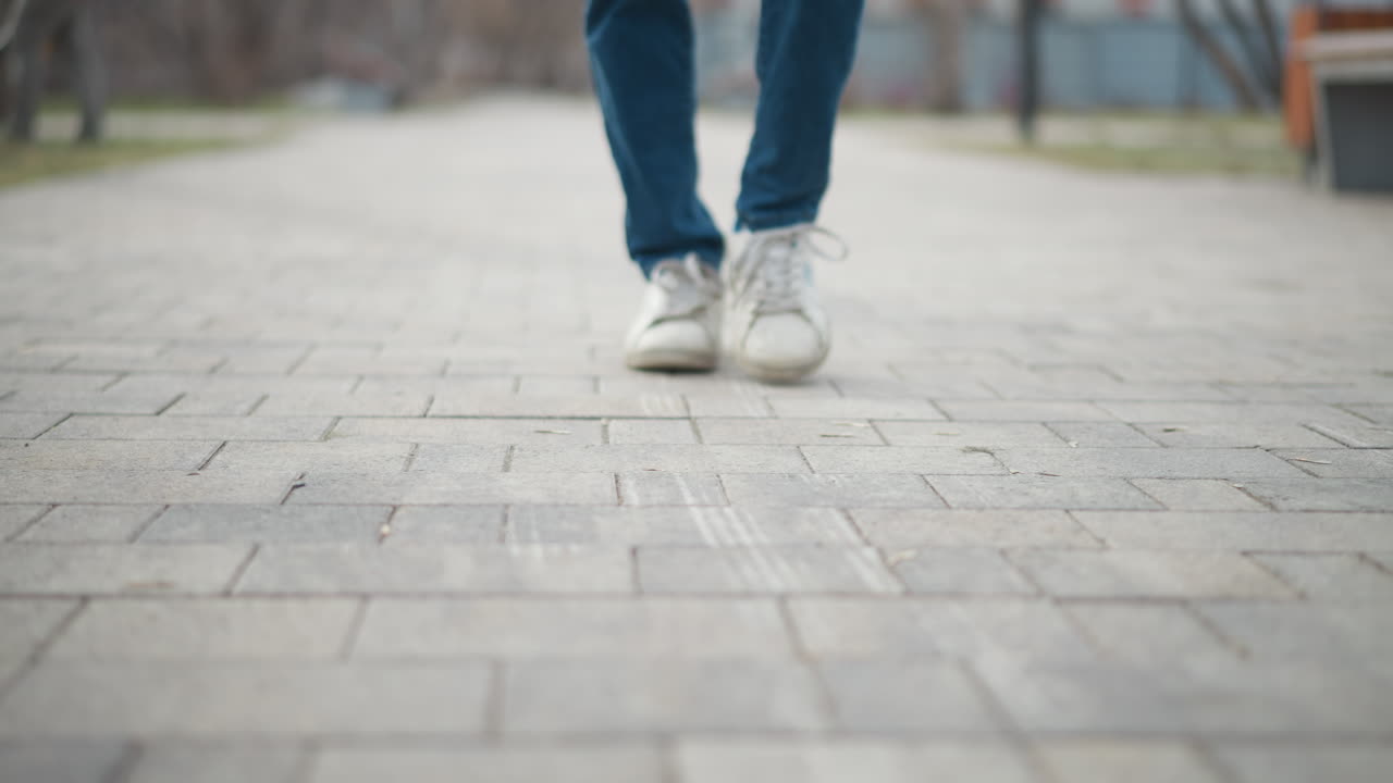 Person walking on paved sidewalk in city park, captured from low angle with motion blur on legs and shoes, emphasizing movement, journey, and passage of time in peaceful outdoor environment