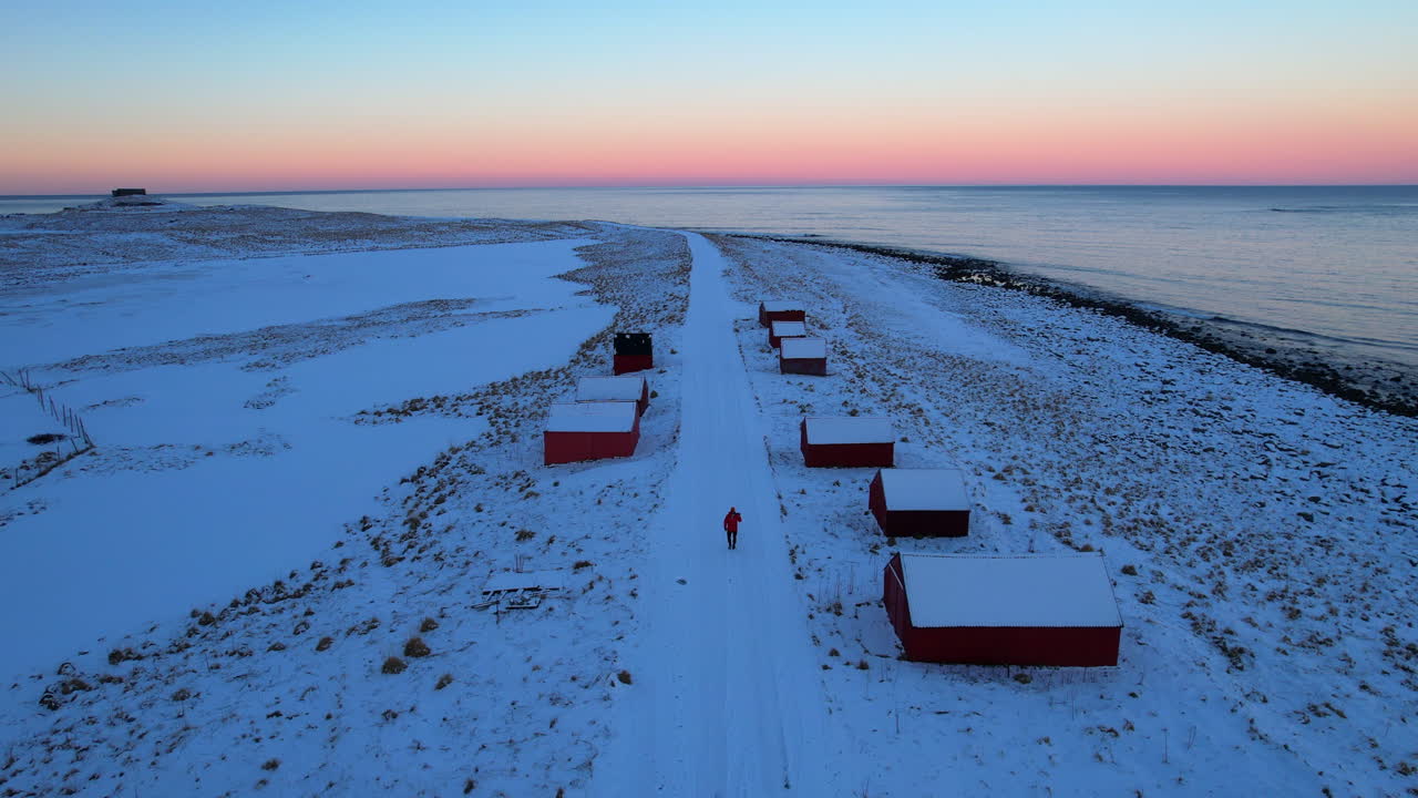 Cinematic 4K drone push in at Eggum. A lonely photographer walks along a snowy coastal road during the blue hour sunrise. Dramatic Arctic sky and frozen landscape in Vestvagoy, Lofoten, Norway