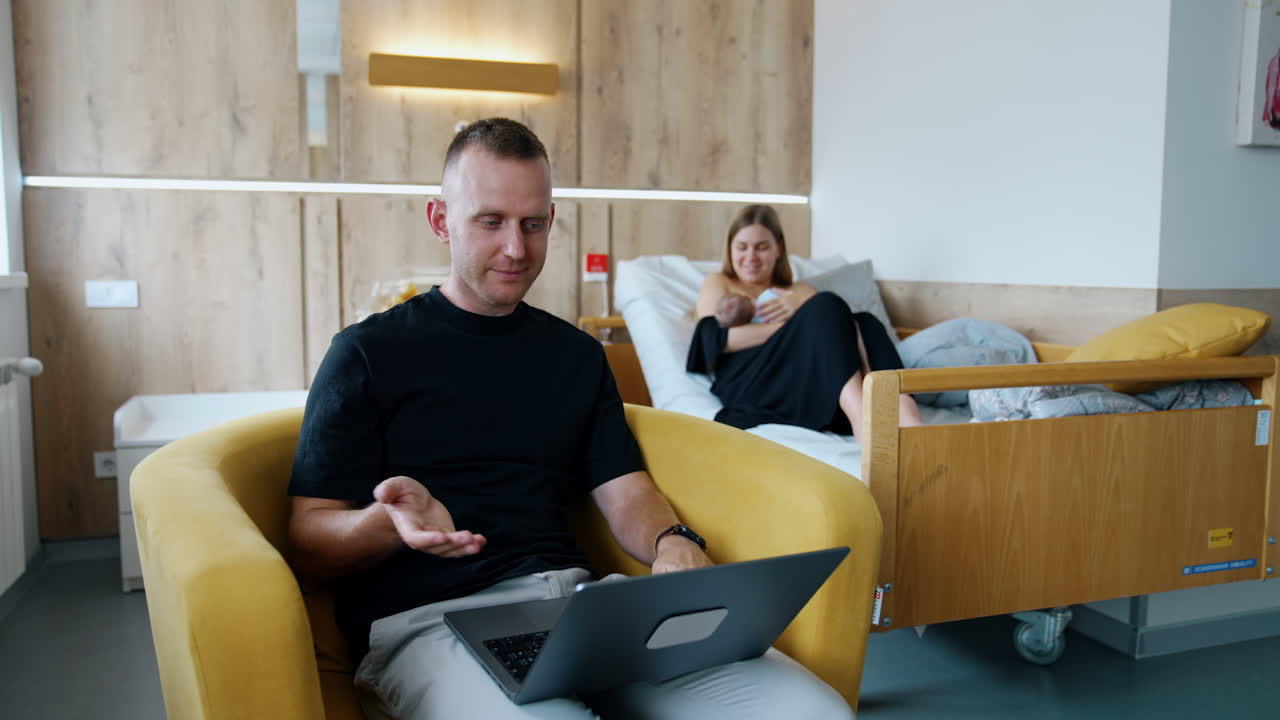 Busy Caucasian man sits in armchair working on laptop. Woman in bed at backdrop breastfeeding her baby.