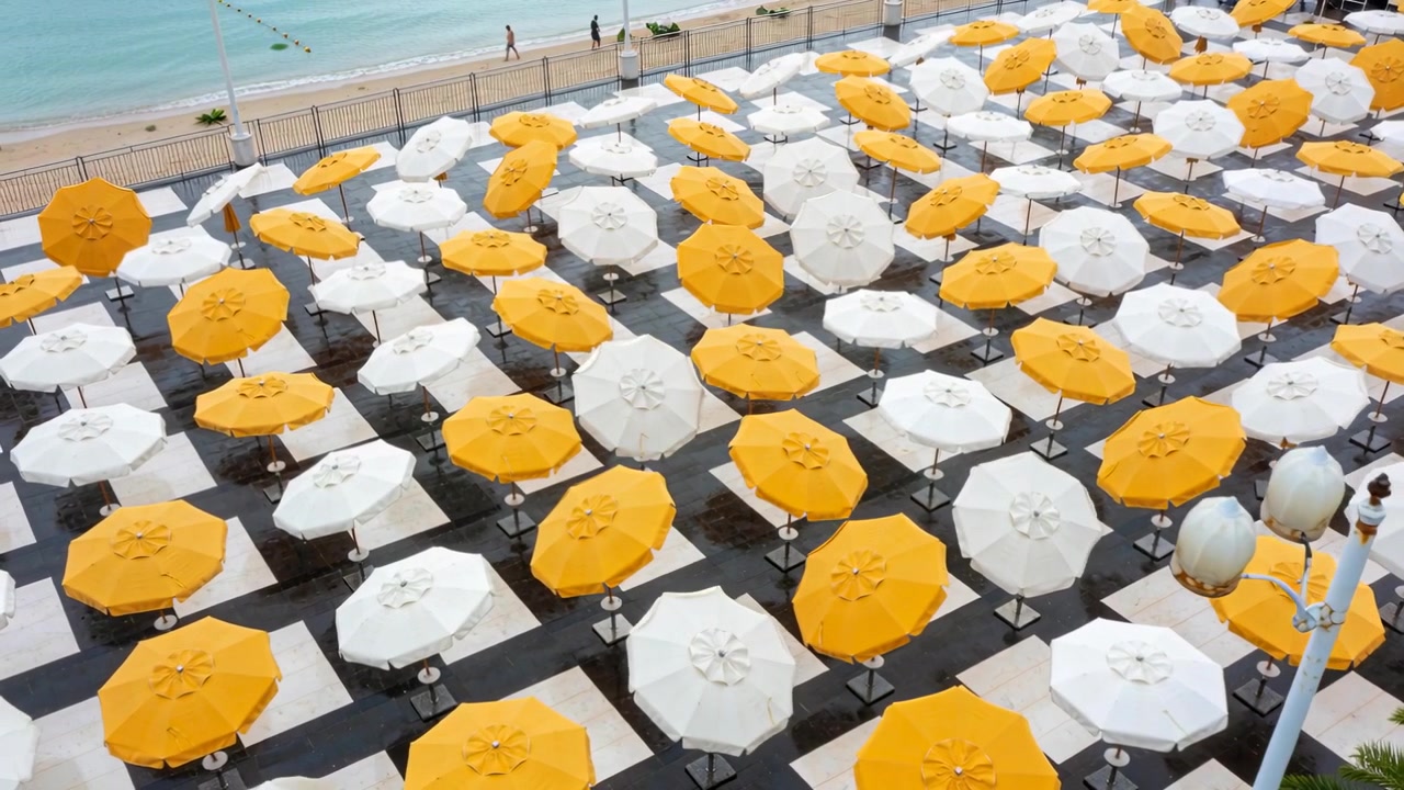 La telecamera di un drone effettua una panoramica rivelando ombrelloni ottagonali su una terrazza sul mare, mentre due persone camminano sul lungomare.