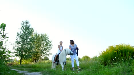 Mom and daughter are walking around the field, daughter is riding a pony, mother is holding a pony for a bridle. Cheerful, happy family vacation. Outdoors, in summer, near the forest