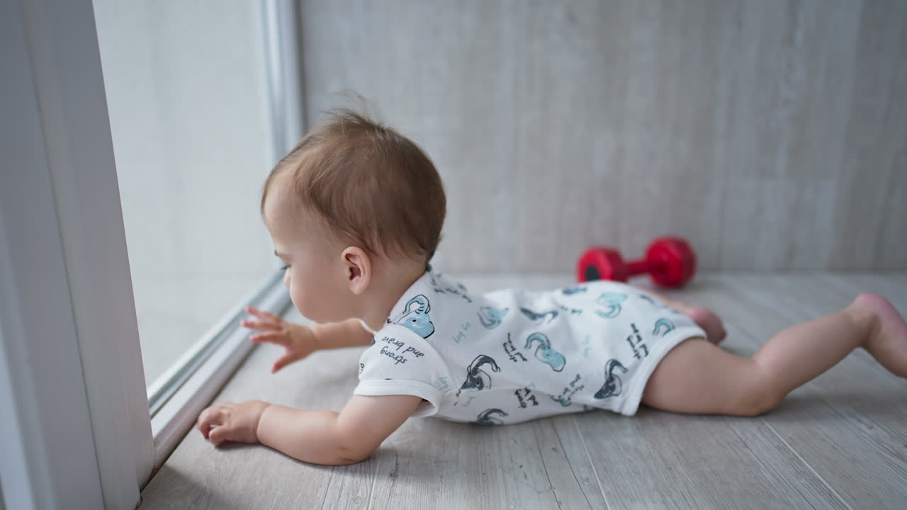 Little cute child on the wooden floor near the glass door. Adorable toddler touching the glass with his hands. Side view.