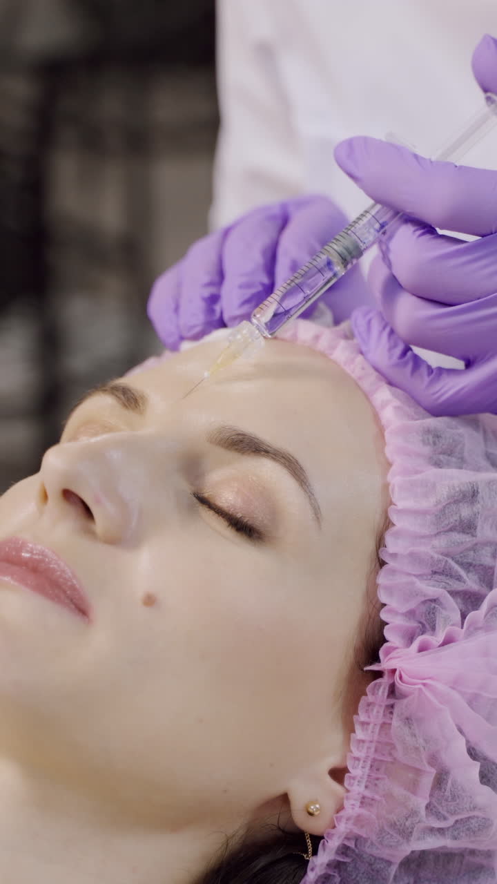 A beautiful woman is on the procedure of facial rejuvenation. Beautician makes mesotherapy of the forehead area. Beauty injections Vertical video