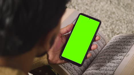 Close-Up-Of-Muslim-Man-At-Home-Sitting-On-Floor-And-Reciting-From-The-Quran-Holding-Green-Screen-Mobile-Phone-1