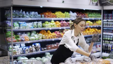 Women-workers-in-black-apron-and-gloves-stocking-the-fruits-in-supermarket.-Young-employee-at-work.-Side