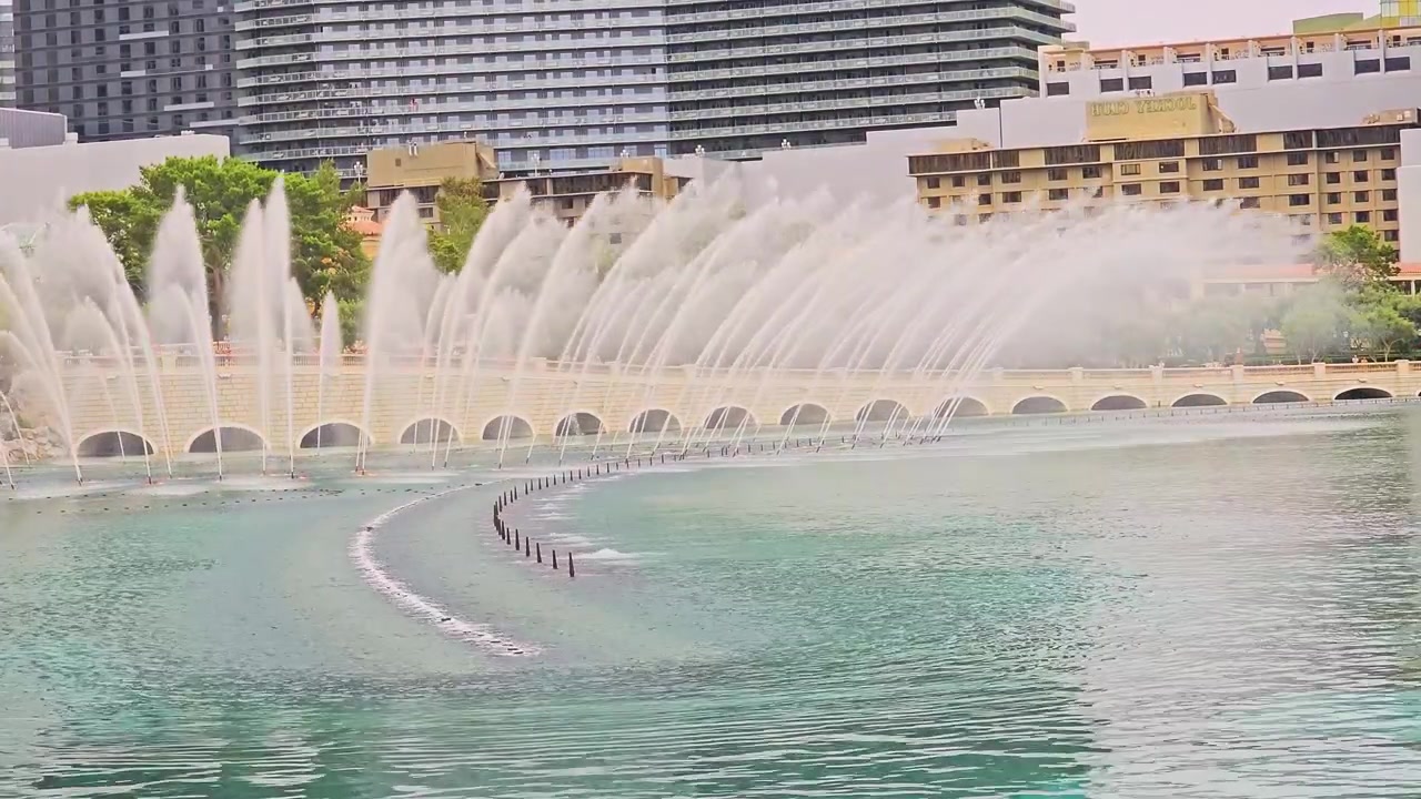 Vista diurna amplia del espectáculo de las fuentes del Bellagio, con altos chorros de agua, ondas turquesas en el lago, el horizonte de hoteles, un muro de piedra arqueado, árboles verdes, turistas y el soleado paisaje del resort de Las Vegas.