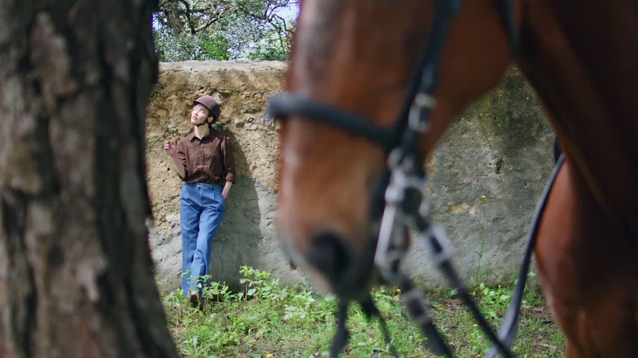 Jockey girl leaning wall at ranch area. Wild brown horse in bridle standing