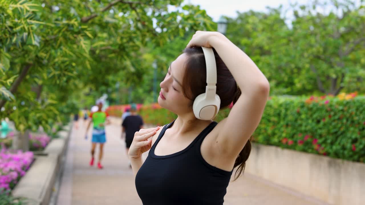 Asian woman performs neck stretches with headphones during morning workout in lush city park