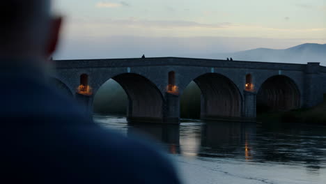 a person views an ancient stone arch bridge over a river at dusk