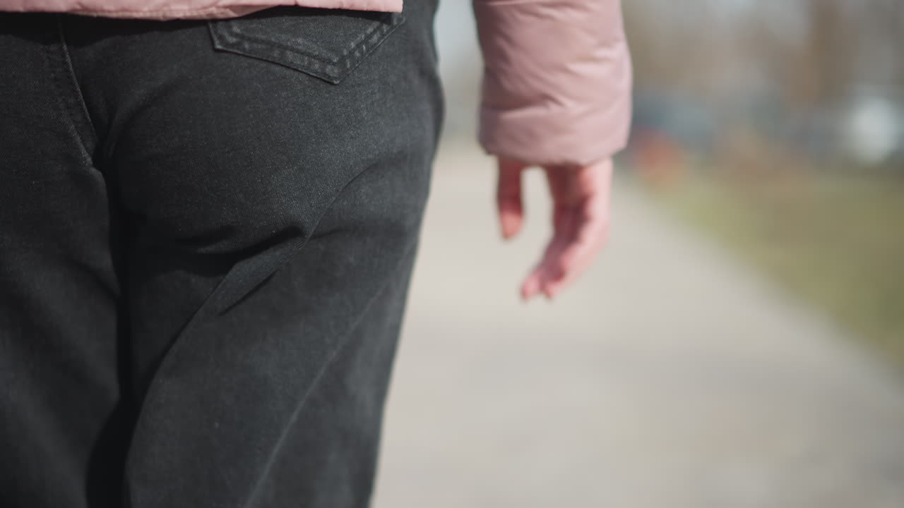 Close-up shot of young woman s lower body from behind, showing pink padded sleeve, relaxed hanging hand, and back pocket of black denim pants while walking outdoors on paved path during cold season