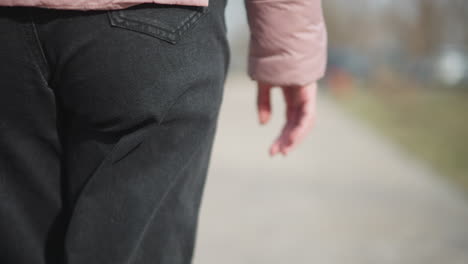 close-up shot of young woman s lower body from behind, showing pink padded sleeve, relaxed hanging hand, and back pocket of black denim pants while walking outdoors on paved path during cold season