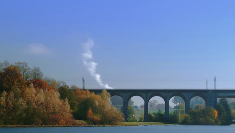 a time-lapse shot of a lake and a railway bridge where people walk underneath and a smoking chimney