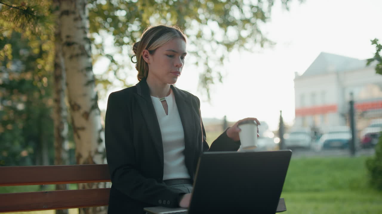 Un'elegante nomade digitale si concentra sul laptop mentre sorseggia l'acqua dalla tazza, seduta su una panchina in un parco soleggiato all'aperto con alberi, un autobus in movimento in lontananza, auto parcheggiate ed edifici urbani