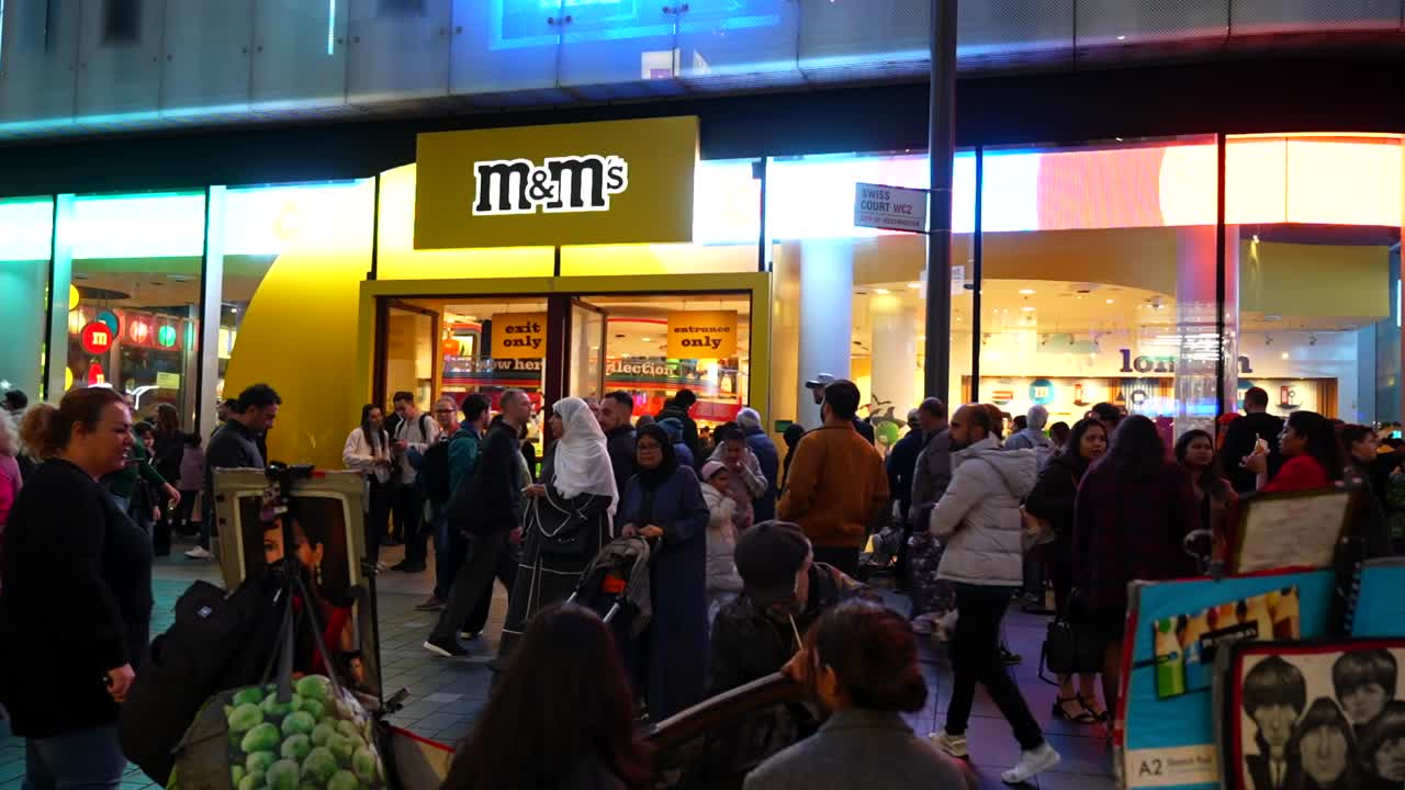 Busy evening scene in Leicester Square with tourists outside M and M's store and a street artist drawing, London