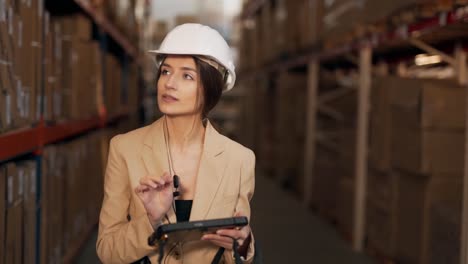 hermosa mujer joven en un traje de negocios con un casco en la cabeza trabajando con una tableta en el fondo del almacén con cajas