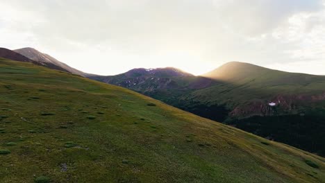 Drone-dolly-above-vibrant-green-hills-reveals-hidden-alpine-lake-below-sun-beam-shooting-through-guanella-pass-colorado-mountains