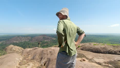 Tiro-En-órbita-En-Cámara-Lenta-Alrededor-De-Un-Hombre-De-Jengibre-Barbudo-Con-Un-Sombrero-Grande-Mientras-Estaba-De-Pie-En-La-Cima-De-Una-Montaña-De-Granito-Y-Mirando-Hacia-Las-Vistas-Del-Este-De-África