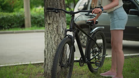 woman in jean shorts approaches bicycle leaning against tree in calm park setting, reaching to remove stand, with green background of trees, paved path, and parked vehicle nearby